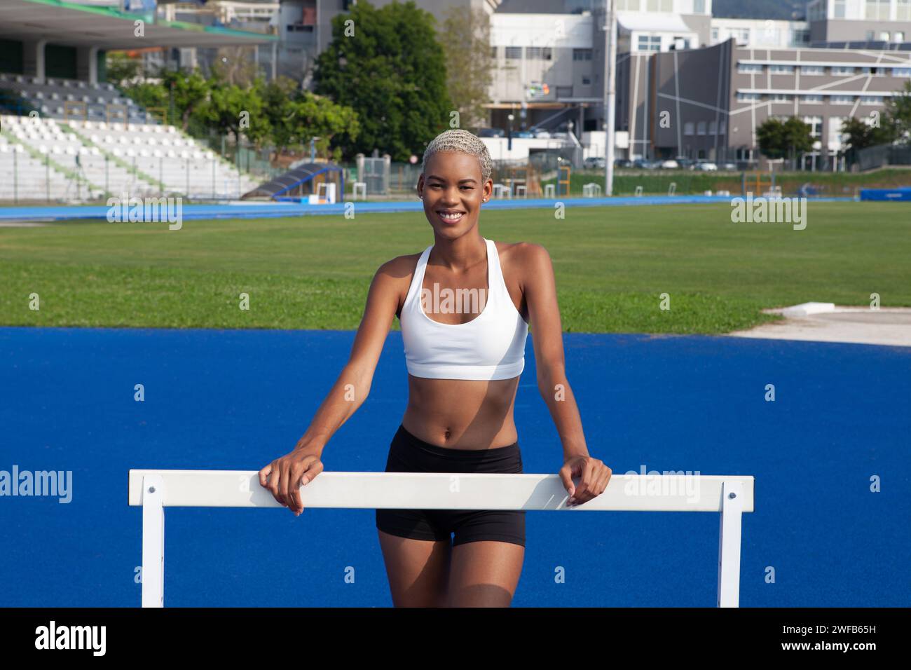 Eine lächelnde afroamerikanische Athletin trainiert auf der Olympiagruppe, lehnt sich an die Hürden, mit blauen Bahnen im Hintergrund, Konzept o Stockfoto