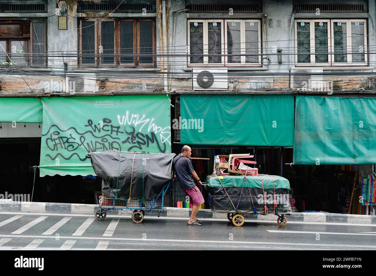 Ein Straßenverkäufer in der Samsen Road, Bangkok, Thailand, hat Schwierigkeiten, seine mobilen Verkaufsstände zu kontrollieren, während er sie auf der abfallenden Straße schiebt/zieht Stockfoto