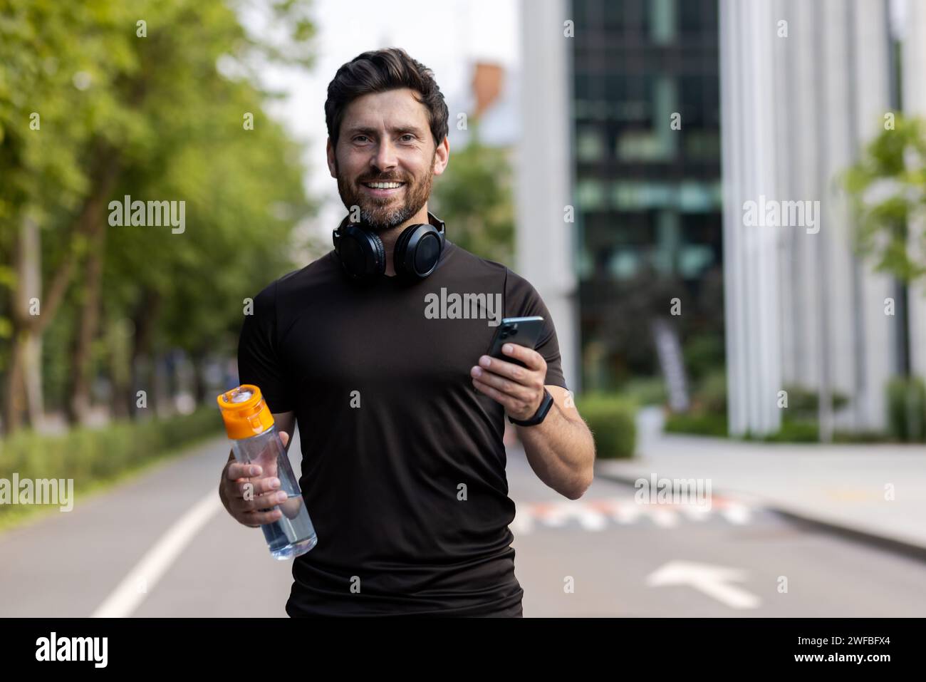 Porträt eines lächelnden jungen Mannes, der auf der Straße der Stadt läuft, in Kopfhörern steht, eine Flasche Wasser und ein Telefon hält und in die Kamera blickt. Stockfoto