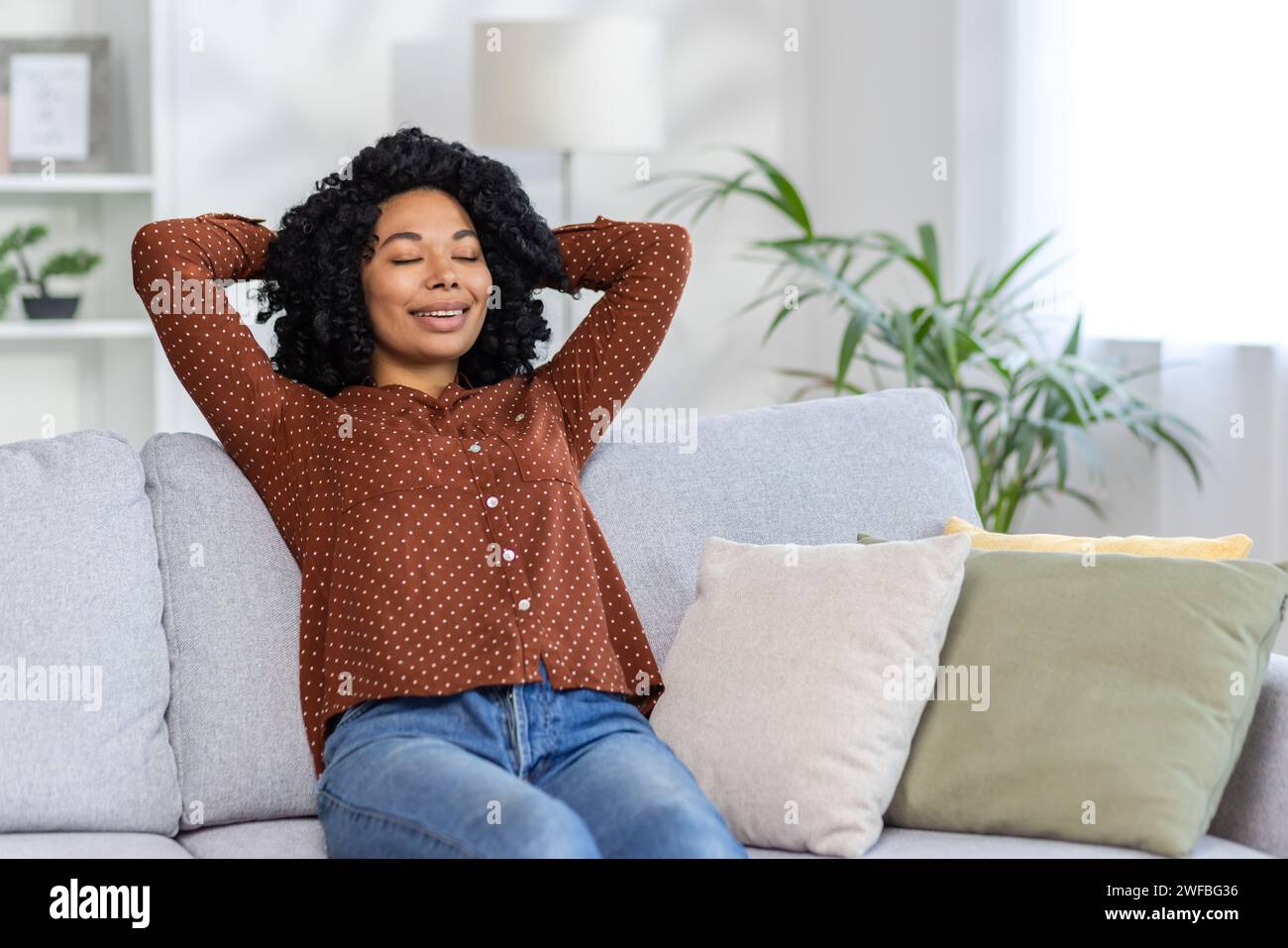 Entspannte afroamerikanische Frau genießt einen friedlichen Moment, liegend auf einer Couch zu Hause, die Augen geschlossen mit einem zufriedenen Lächeln. Stockfoto