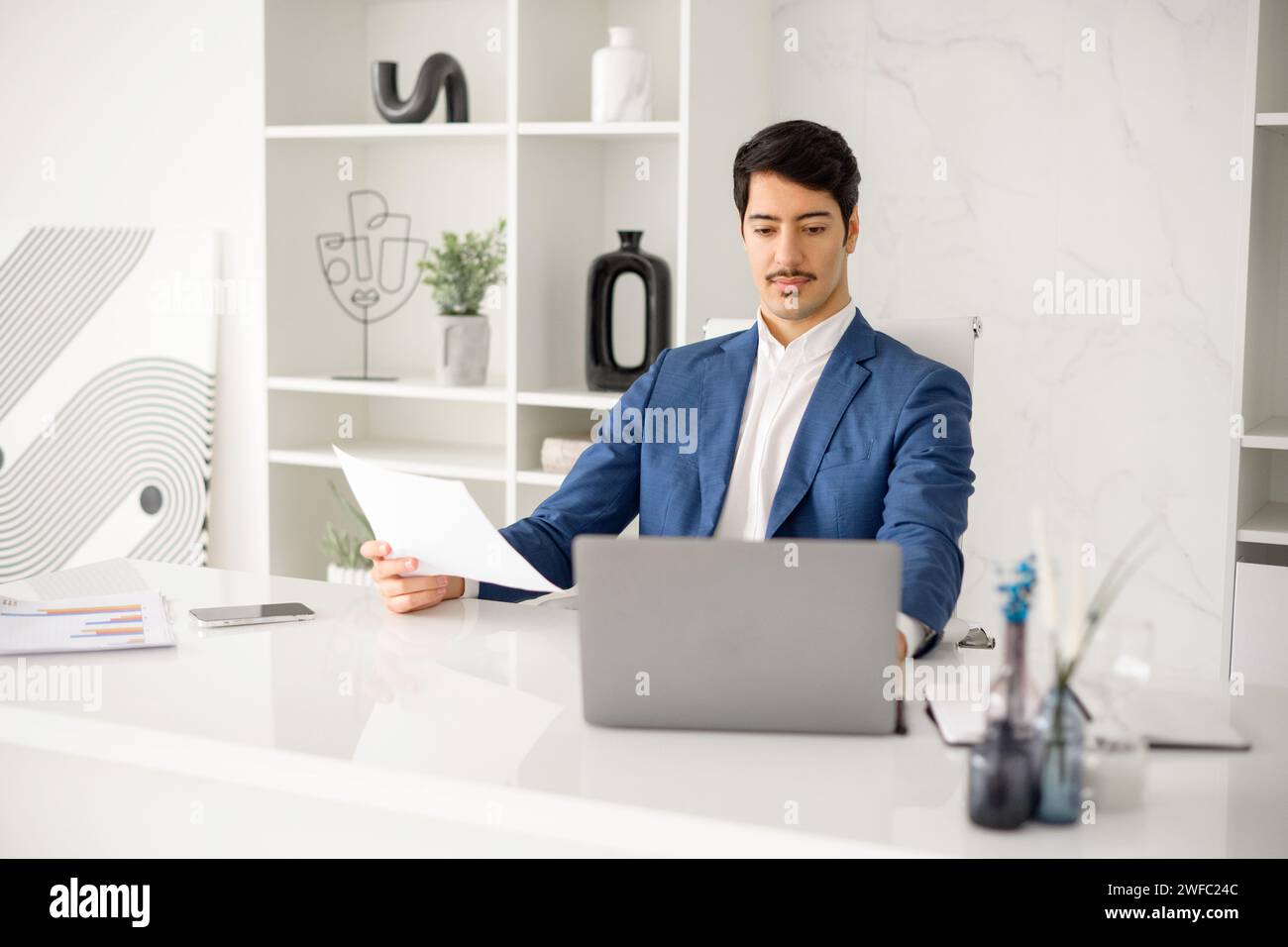 Ein fleißiger hispanischer Geschäftsmann ist in der Lektüre eines Papierdokuments vertieft, während er Aufgaben auf seinem Laptop verwaltet, was die Multitasking-Anforderungen aktueller Unternehmensrollen veranschaulicht Stockfoto