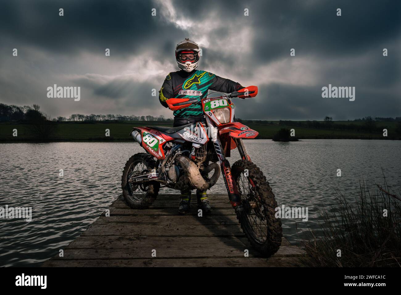 Der Fahrer stand mit einem Off-Road-Motorrad auf einem Holzpier über einem See mit einem stürmischen Himmel im Hintergrund Stockfoto