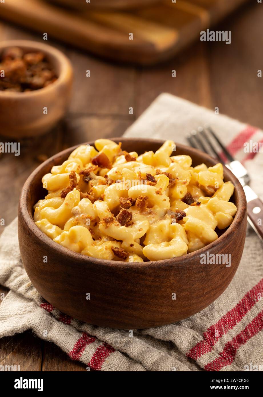 Makkaroni und Käse mit Speckstücken in einer Holzschale auf einem Handtuch und rustikalem Holztisch Stockfoto