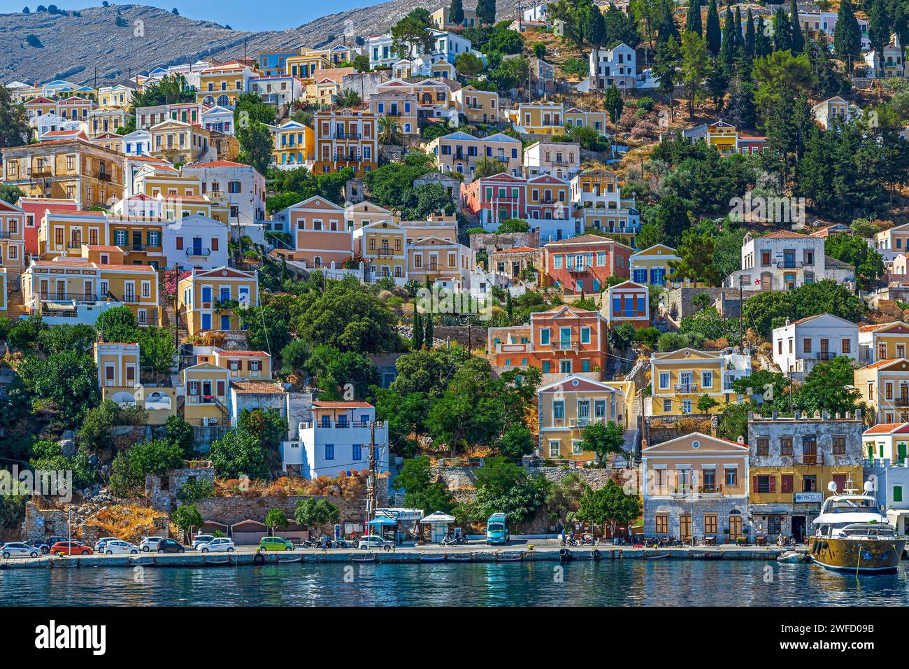 SYMI, GRIECHENLAND - 3. JULI 2022: Hafen von Symi mit farbenfrohen neoklassizistischen Herrenhäusern, die die Hänge in der Nähe der Stadt bedecken. Teil der Dodekanesischen Insel. Stockfoto