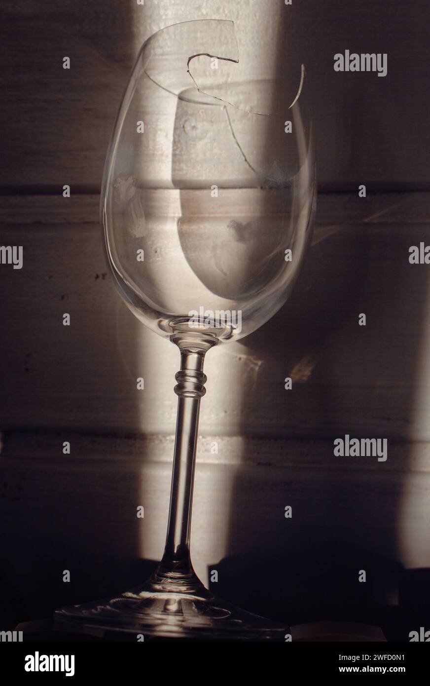 Zerbrochenes Weinglas im Sonnenlicht auf hölzernem Hintergrund. Beschädigtes Weinglas mit scharfen Kanten. Zerrissenes Glas im Schatten. Kreatives Stillleben. Zerbrechliches Objekt Stockfoto