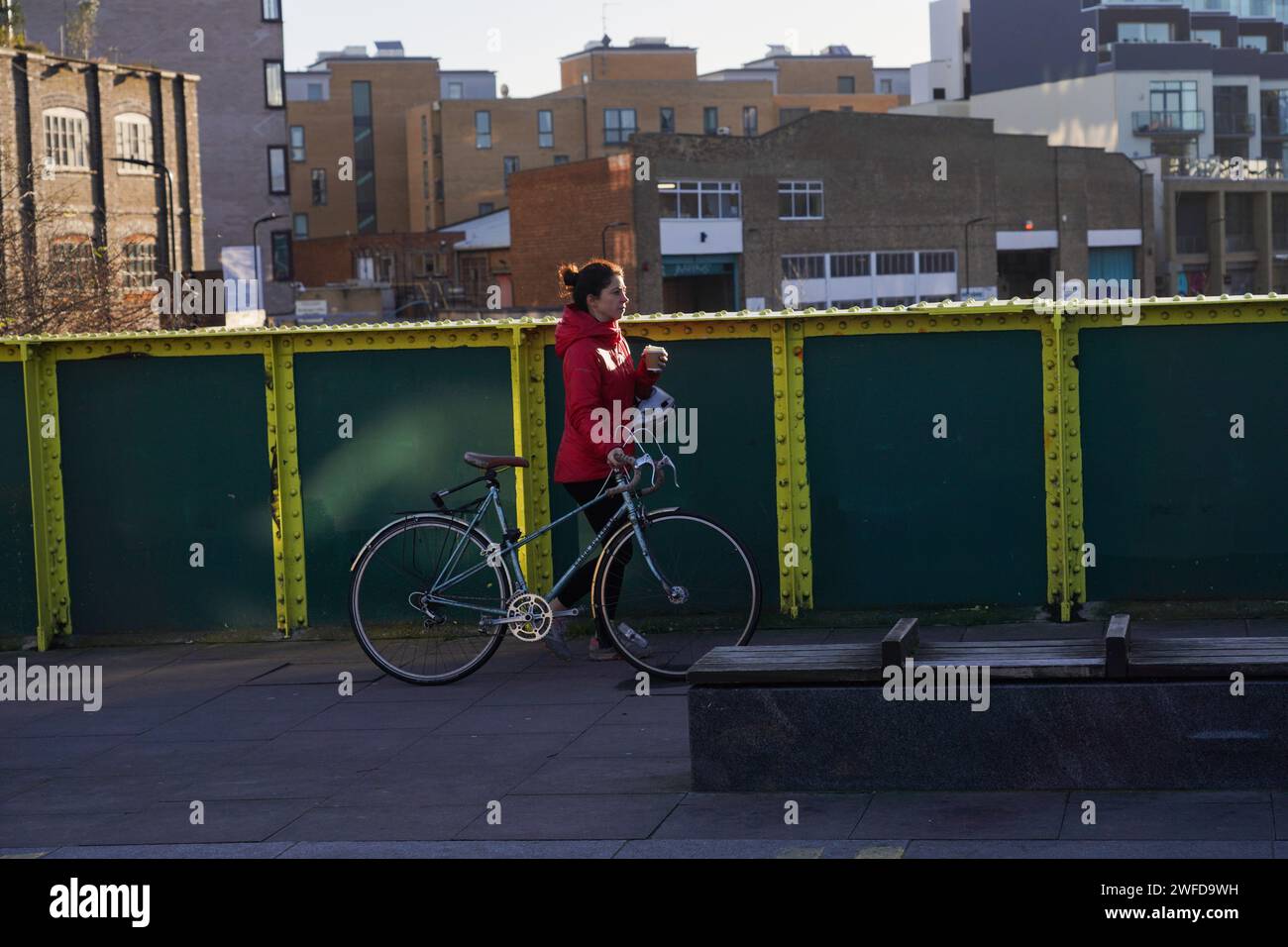 Eine weiße Fahrradfahrerin läuft mit ihrem Kaffee oder Tee zum Mitnehmen und zieht ihr Fahrrad in der frühen Morgensonne. Stockfoto
