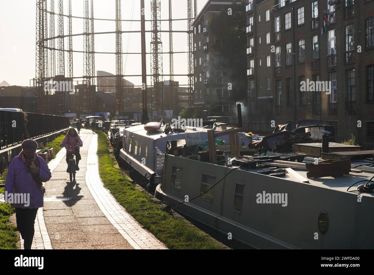 Ein Radfahrer pendelt in der frühen Morgensonne am Regent's Canal entlang. Hausboote sind auf der ganzen Seite des Kanals verankert, wo die Menschen leben. Stockfoto