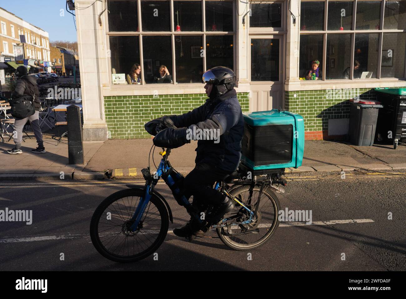 Ein Deliveroo-Mann fährt sein Fahrrad in der frühen Wintersonne am Broadway Market in Hackney, London, Großbritannien. Stockfoto