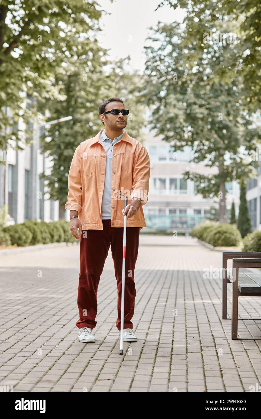 Hübscher indischer Blinder mit Brille und Gehstock in leuchtend orangefarbener Jacke, der im Park spaziert Stockfoto