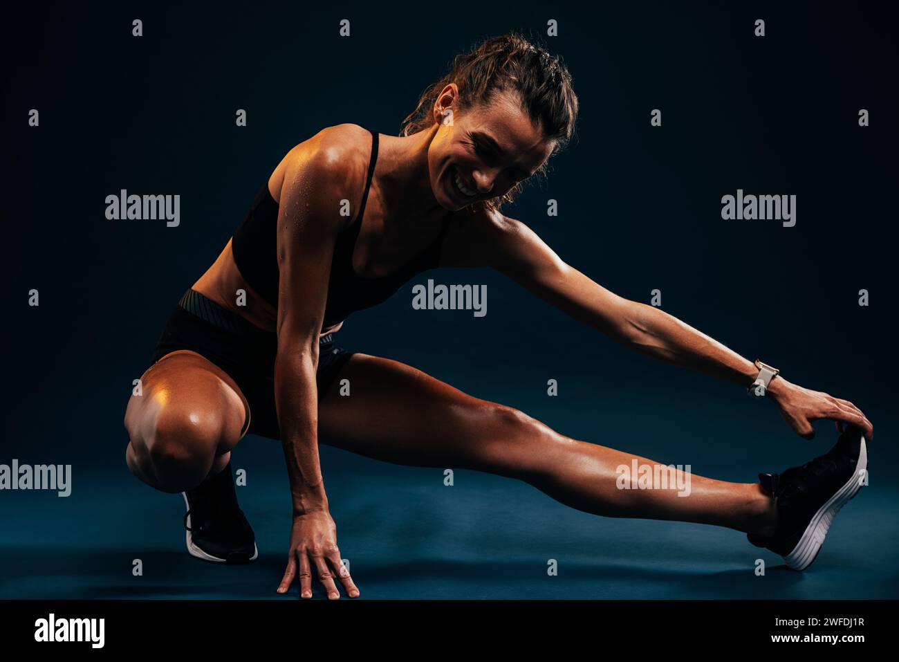 Die junge Athletin lächelt, während sie ihre Beine auf blauem Hintergrund ausdehnt, ein leiser Blitz Stockfoto