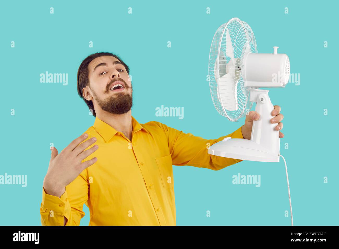 Mann mit Hilfe der kalten Luft vom elektrischen Ventilator entweicht aus der Sommerhitze auf türkisfarbenem Hintergrund. Stockfoto