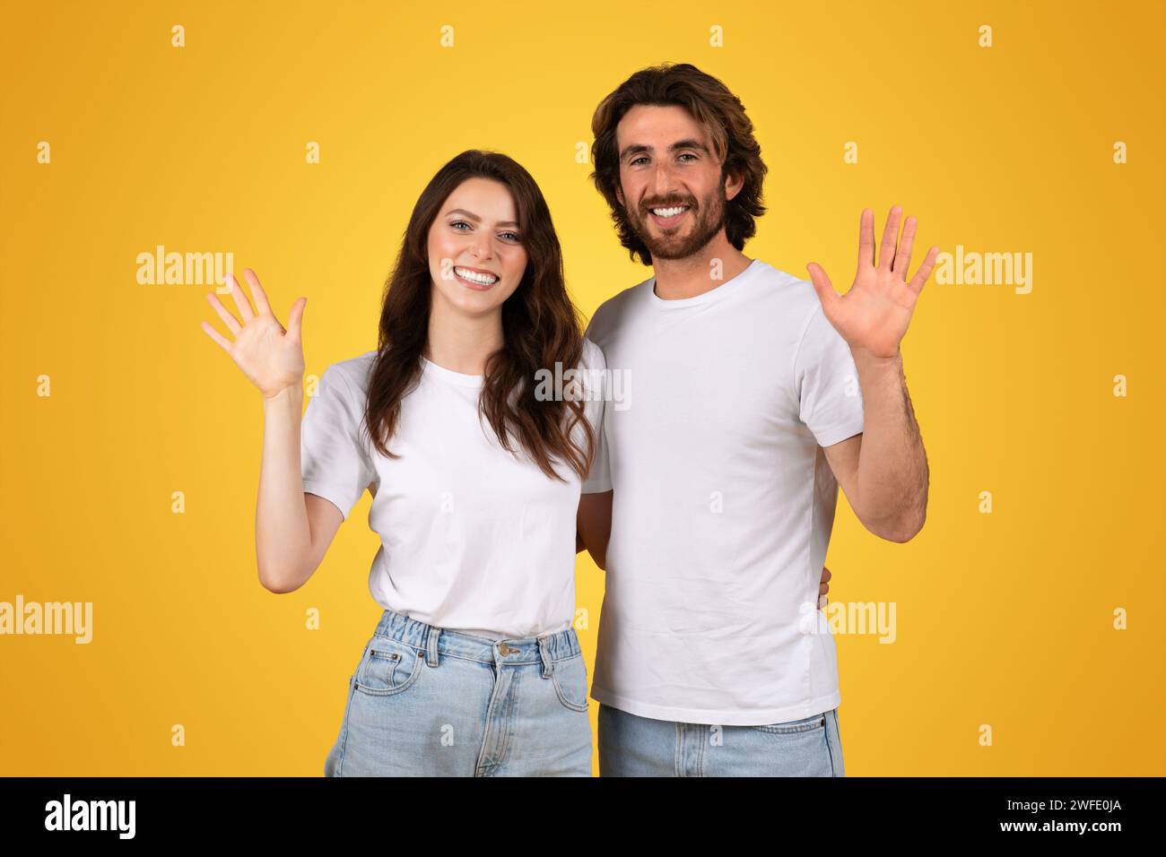 Lächelndes junges Paar in weißen Hemden, das mit einer geöffneten Handgeste hallo winkt Stockfoto