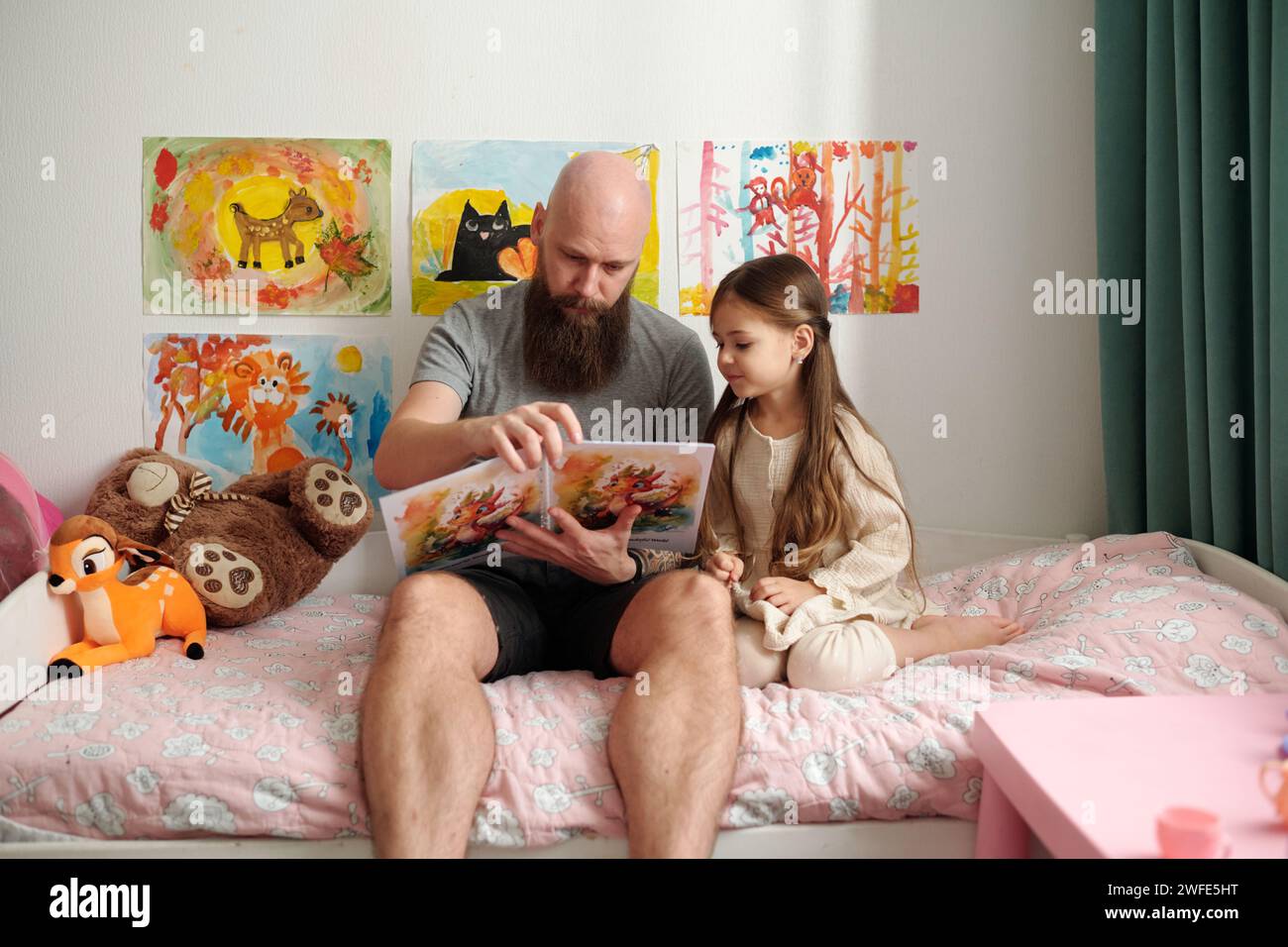 Bärtiger Mann mit offenem Comicbuch mit Bildern, die neben seiner liebenswerten kleinen Tochter auf dem Bett sitzen und ihre neuen Geschichten lesen Stockfoto