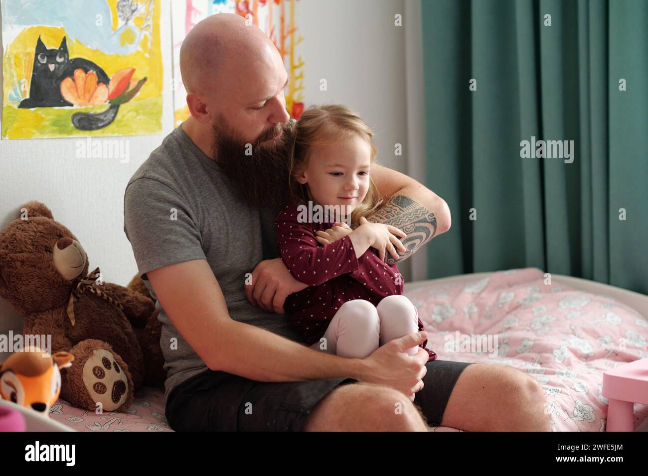 Junger bärtiger Mann im grauen T-Shirt, der die süße blonde Tochter auf den Knien hält und sie umschließt, während beide mit Spielzeug auf dem Bett sitzen Stockfoto