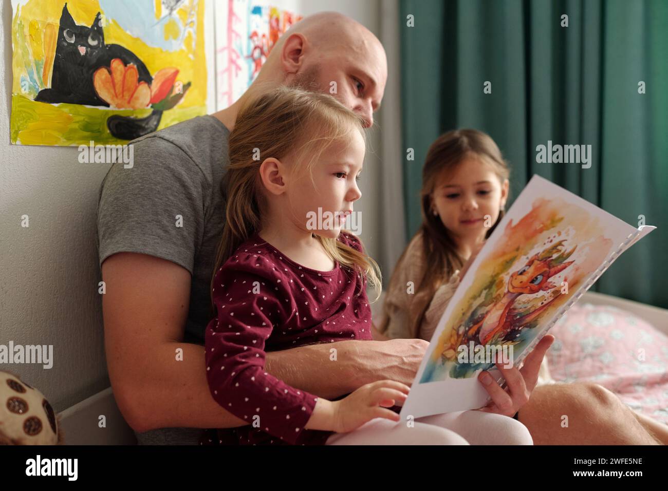 Der liebevolle Vater liest ein Buch mit Bildern für seine süßen kleinen Töchter, während er vor der Kamera sitzt und eine von ihnen umschließt Stockfoto