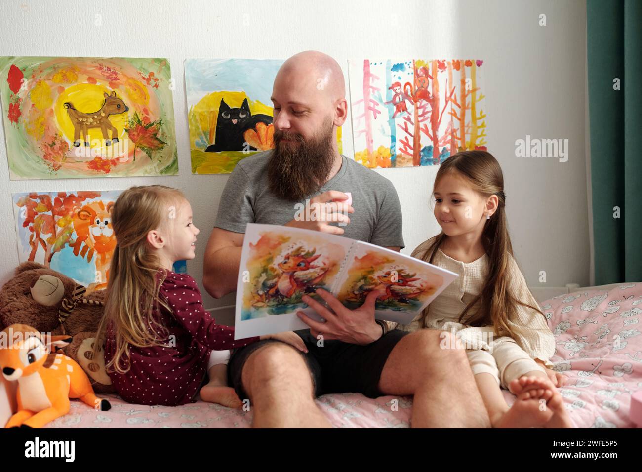Glücklicher bärtiger Mann mit offenem Buch, der eine seiner süßen kleinen Töchter ansieht, während er zwischen ihnen auf dem Bett sitzt und Comics liest Stockfoto