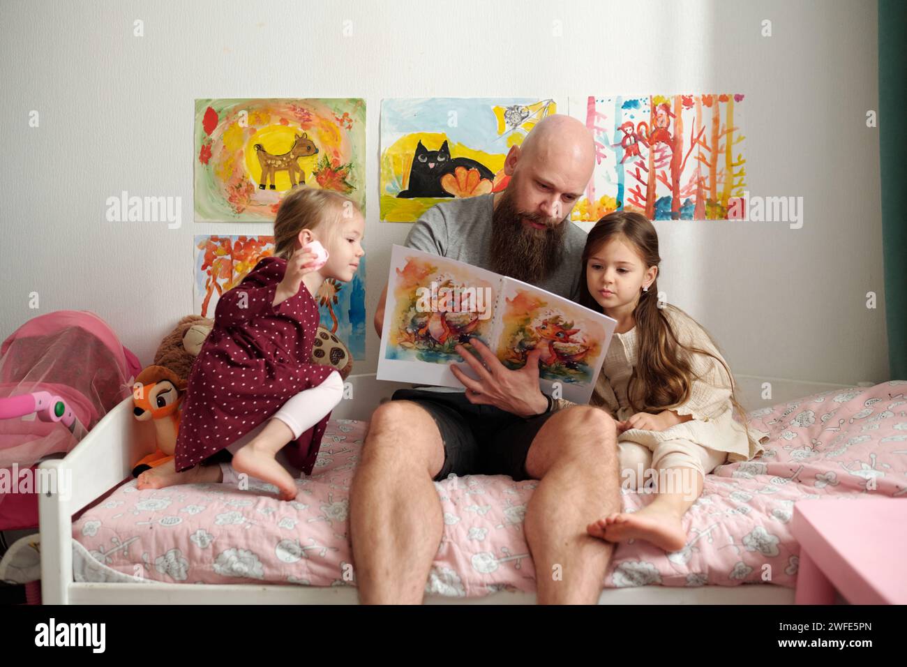 Bärtiger Mann mit einem Buch mit Comics, das Bilder einer seiner süßen kleinen Töchter zeigt, während er neben dem Mädchen in weißem Kleid auf dem Bett sitzt Stockfoto