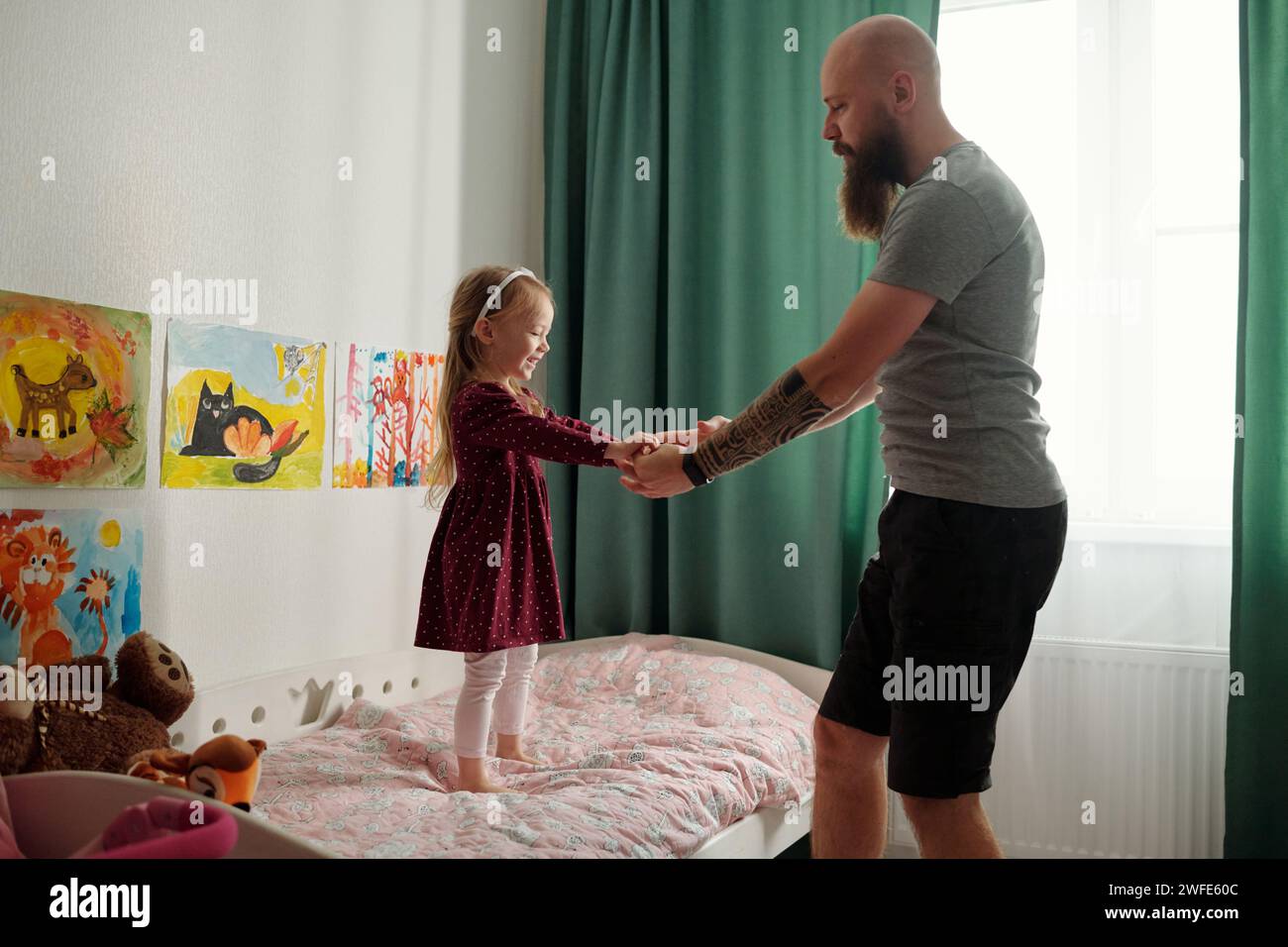 Seitenansicht eines jungen bärtigen Vaters, der seine liebenswerte kleine Tochter an den Händen hält, während er vor dem Bett vor grünen Vorhängen steht Stockfoto