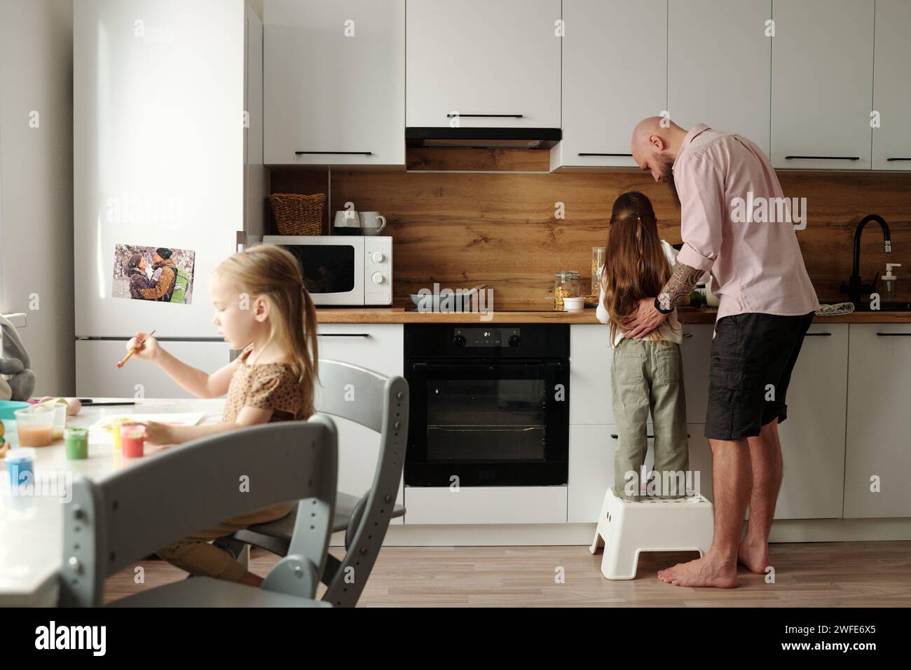 Rückansicht eines jungen Mannes, der sich über eine seiner süßen kleinen Töchter beugt und ihm dabei hilft, morgens an der Küchentheke das Frühstück zuzubereiten Stockfoto