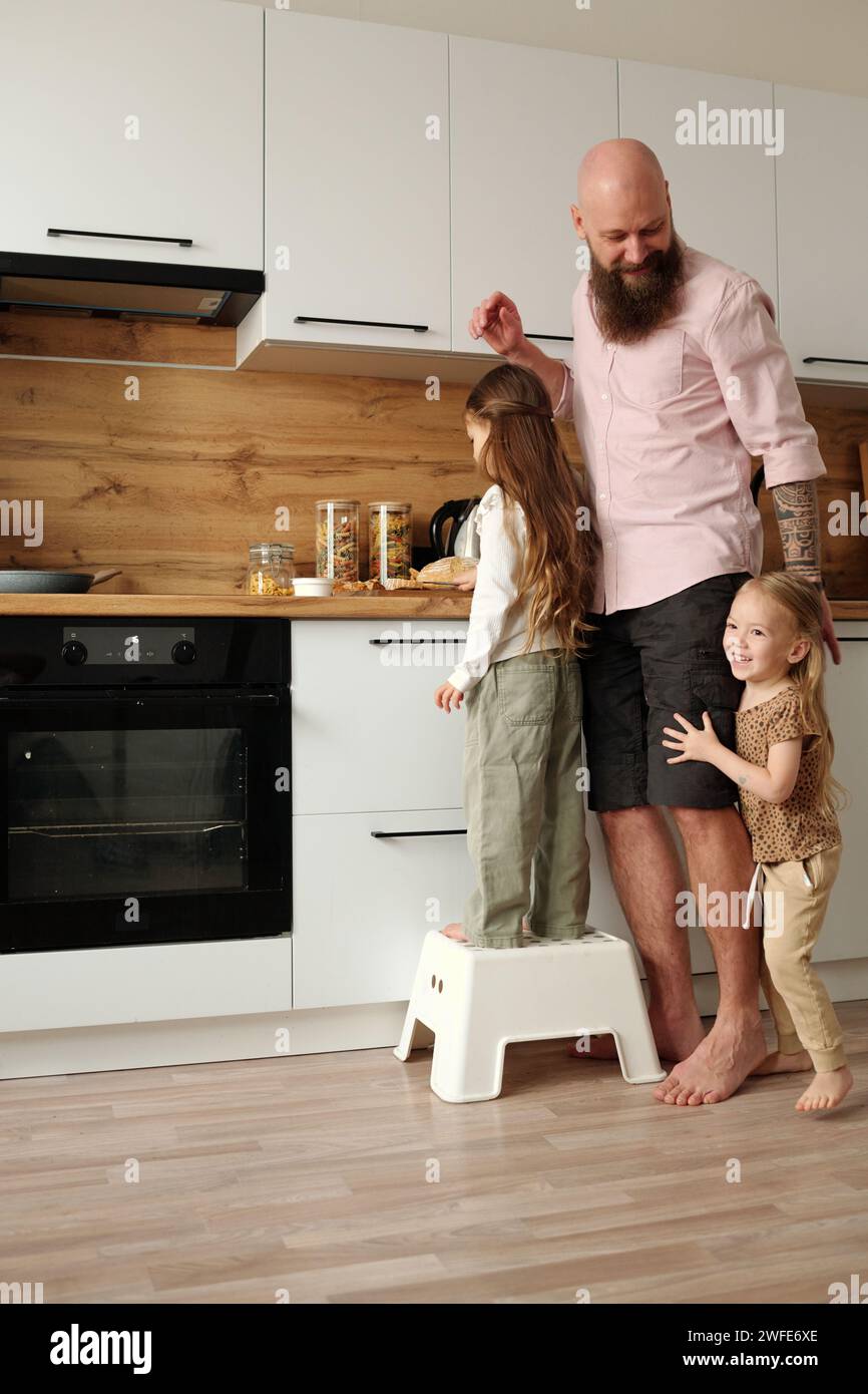 Süßes, fröhliches Mädchen, das Spaß neben ihrem Vater hat, neben einer anderen Tochter zu stehen und ihm dabei hilft, einen Snack für das Frühstück an der Küchentheke zuzubereiten Stockfoto