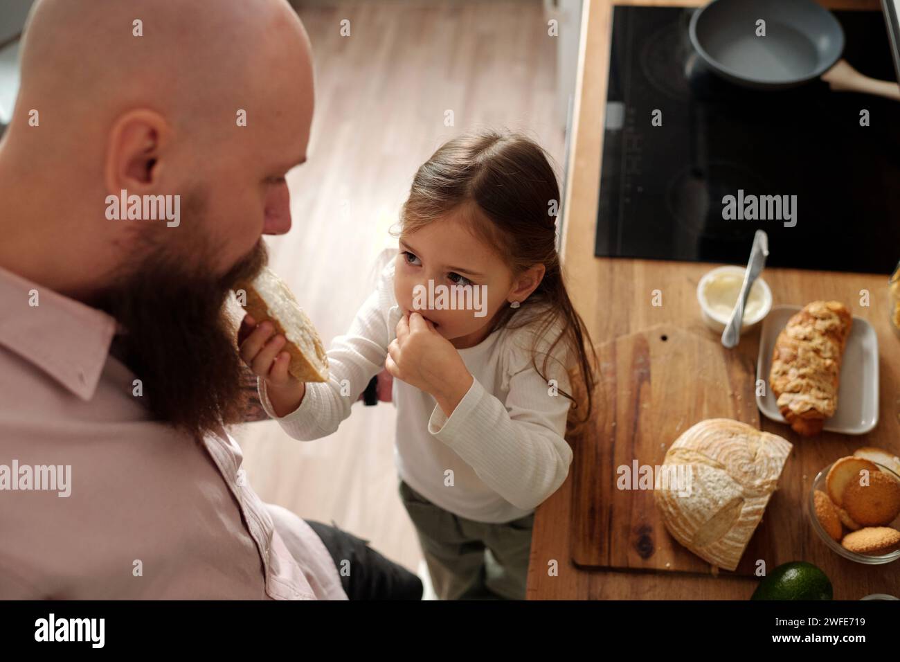 Der hohe Winkel eines süßen Mädchens in weißem Pullover, der ein Sandwich in den Mund seines bärtigen Vaters legt, der während des Frühstücks vor ihr steht Stockfoto