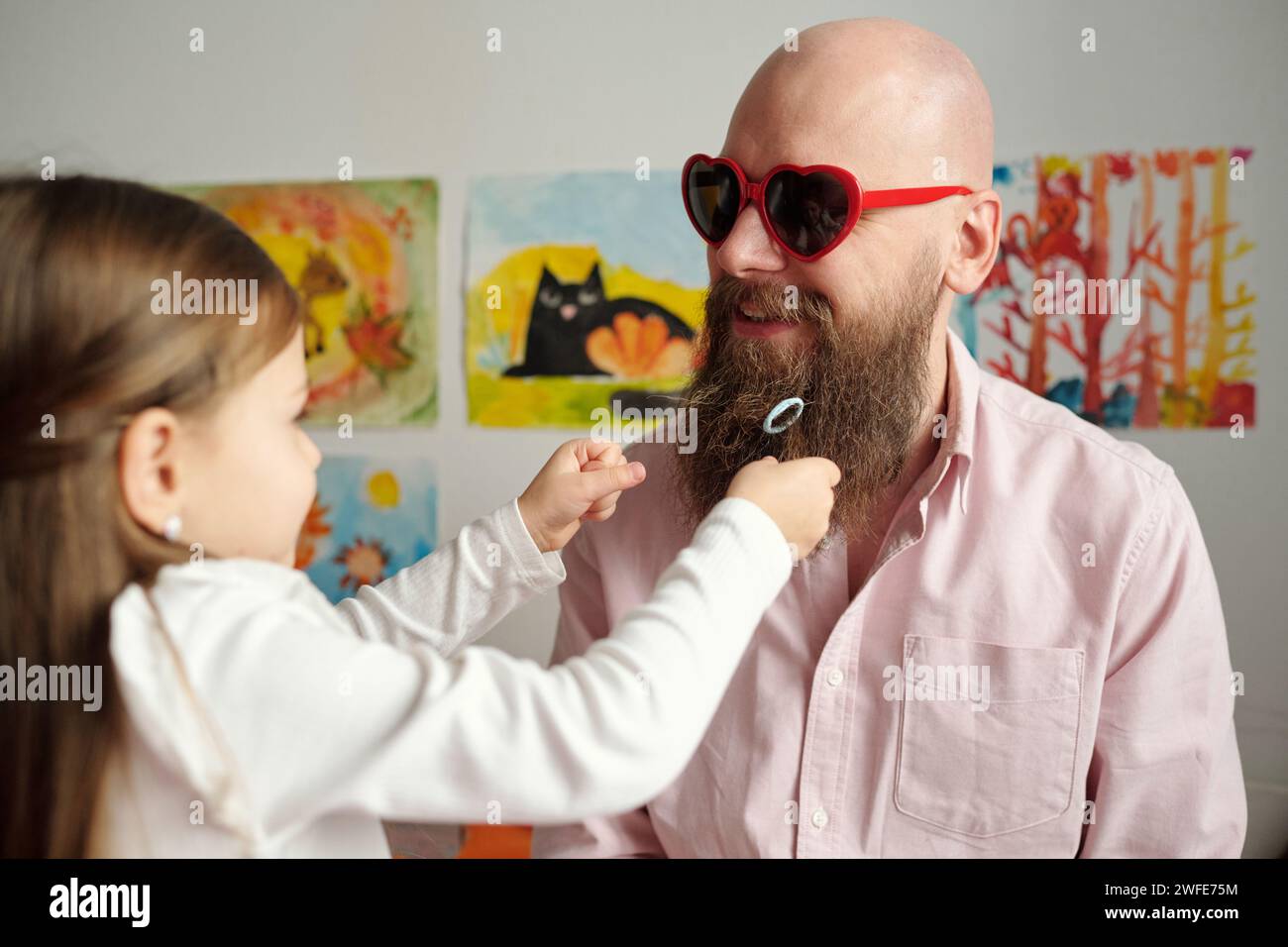 Ein kleines Mädchen, das mit dickem Bart ihres glücklichen Glatzenvaters spielt, in einer herzförmigen Sonnenbrille und einem Hemd, das vor seiner Tochter steht Stockfoto