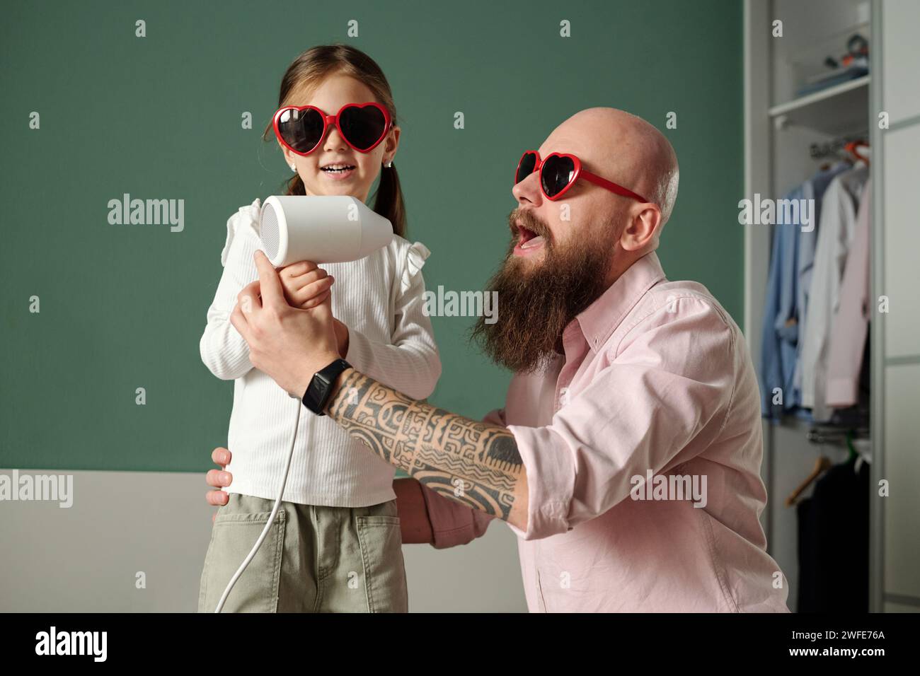 Niedliches kleines Mädchen und ihr kahlköpfiger bärtiger Vater in stilvoller herzförmiger Sonnenbrille singen im Haartrockner, während sie zu Hause Spaß haben Stockfoto