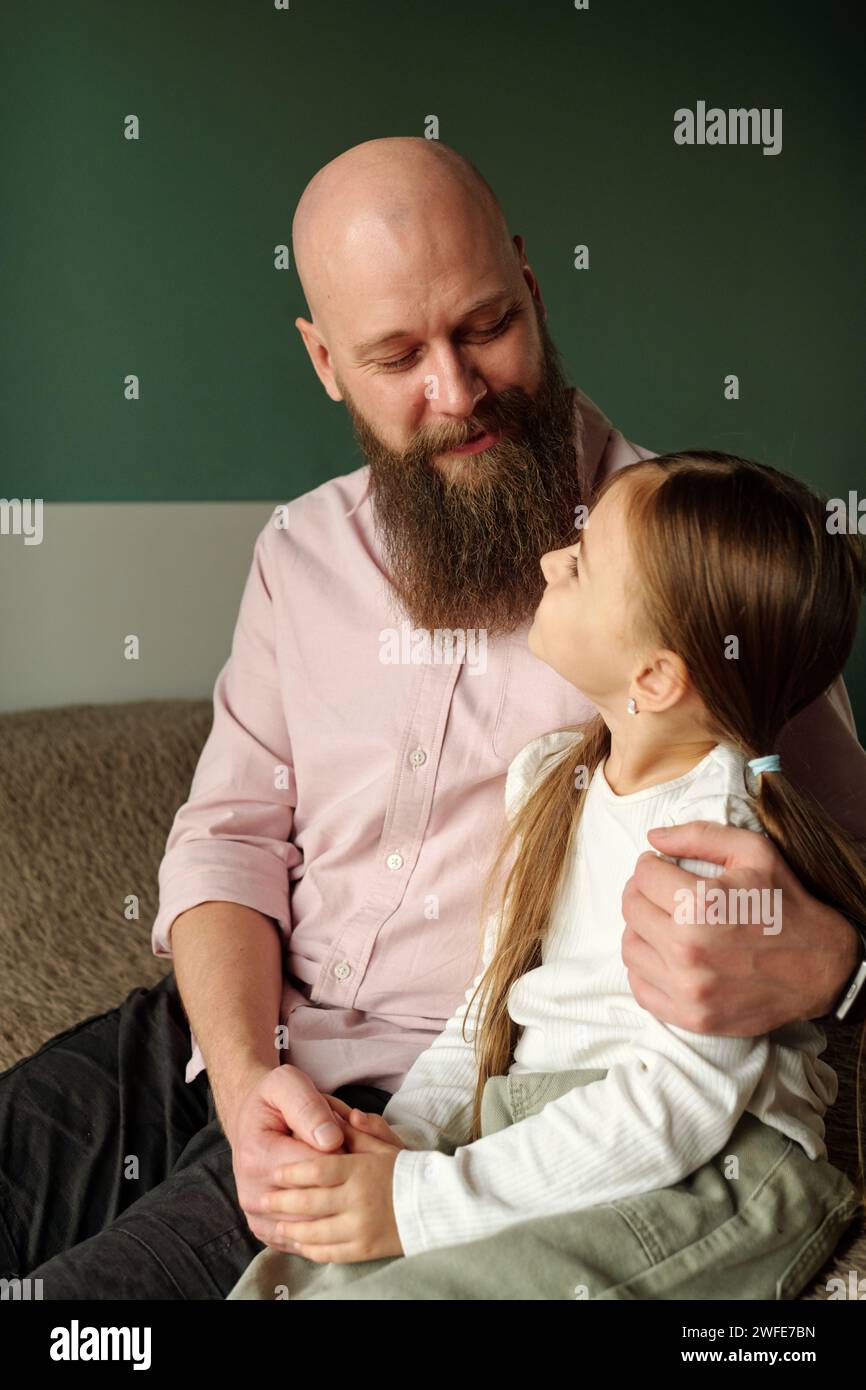 Glücklicher junger bärtiger Mann, der seine entzückende kleine Tochter ansieht und sie umarmen kann, während beide zu Hause vor der Kamera sitzen Stockfoto