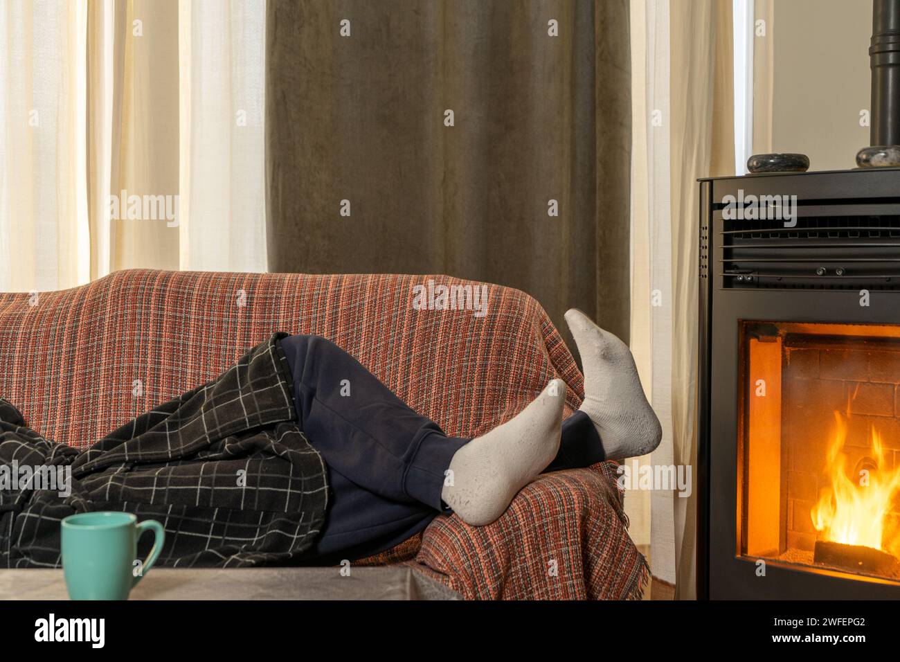 Die Füße des Mannes mit Socken in der Nähe eines Pelletofens, um warm zu bleiben, der Mann liegt auf dem Sofa neben dem Herd, um dem kalten Winter besser standzuhalten. Stockfoto