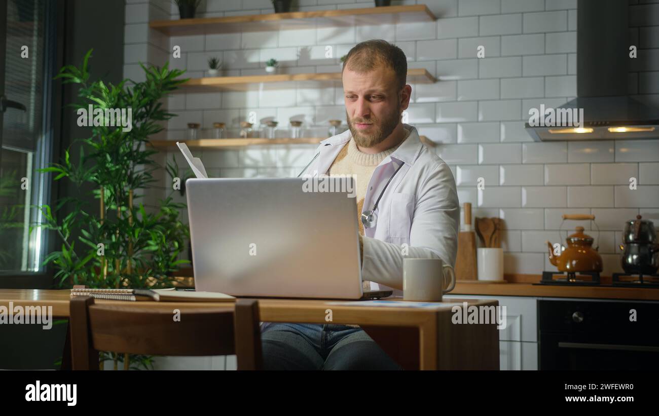 Erfahrener Arzt im weißen Mantel mit Laptop-Computer, der Patientenberichte im Heimbüro untersucht. Männlicher Praktizierender, der Papierkram macht Stockfoto
