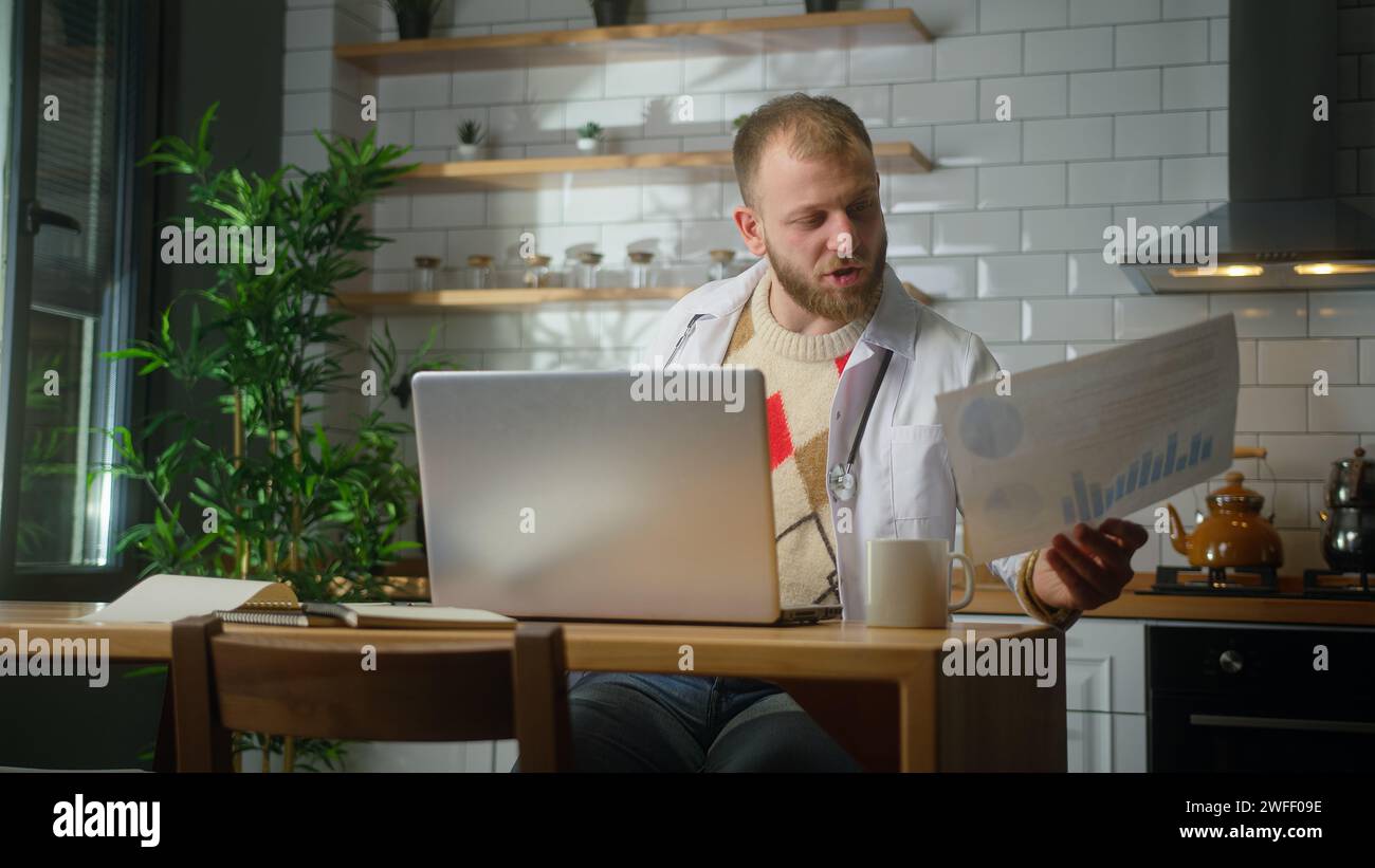 Beschäftigter Arzt mit weißem Mantel mit Laptop-Computer, der Patientenberichte im Heimbüro studiert. Männlicher Praktizierender, der Papierkram macht Stockfoto