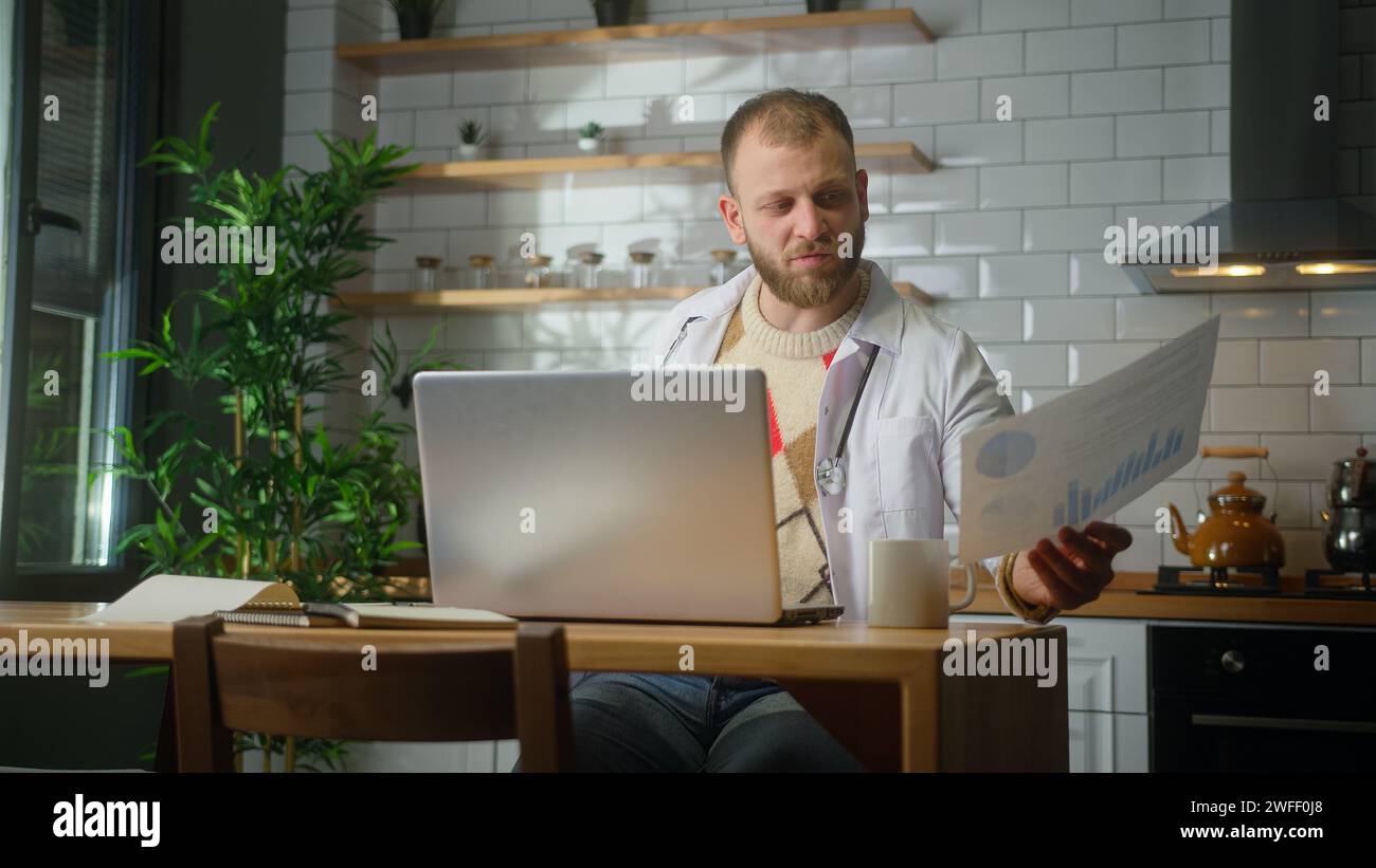Männlicher Arzt mit Laptop-Computer, der Patientenberichte im Heimbüro untersucht. Männlicher Praktizierender, der Papierkram macht Stockfoto