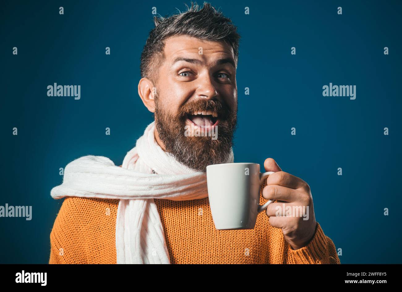 Glücklicher bärtiger Mann in orangefarbenem Pullover und weißem Schal genießt eine Tasse Kaffee oder Tee im Café. Nahaufnahme Porträt eines gutaussehenden Mannes in modischer Kleidung Stockfoto