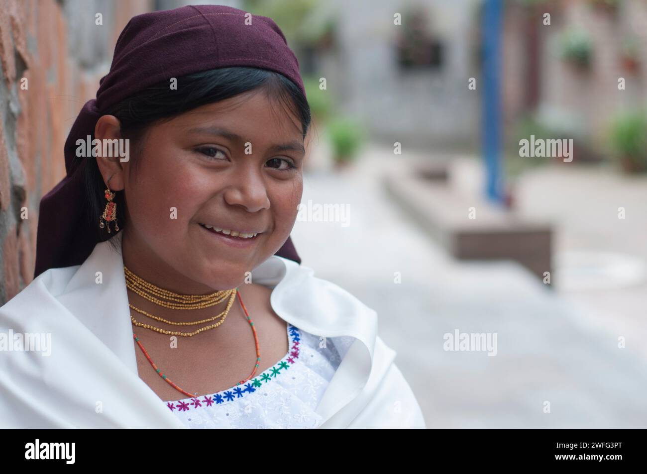 Ein junges indigenes Mädchen in traditioneller Kleidung mit Kopftuch und Schmuck lächelt warm, während es sich an eine Backsteinmauer in einem Kopfsteinpflaster lehnt Stockfoto