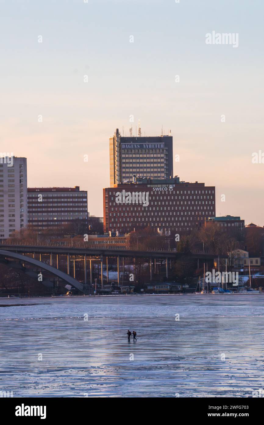 Zwei Personen laufen auf Eis auf dem Mälaren-See in Stockholm, in der Nähe von Kungsholmen und den Gebäuden DN und Aftonbladet. Winter, früher Sonnenuntergang. Klarer Himmel. Stockfoto