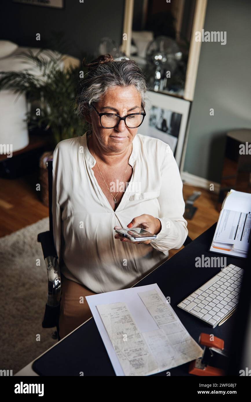 Geschäftsfrau mit Behinderung fotografiert Dokumente, während sie im Heimbüro am Schreibtisch sitzt Stockfoto