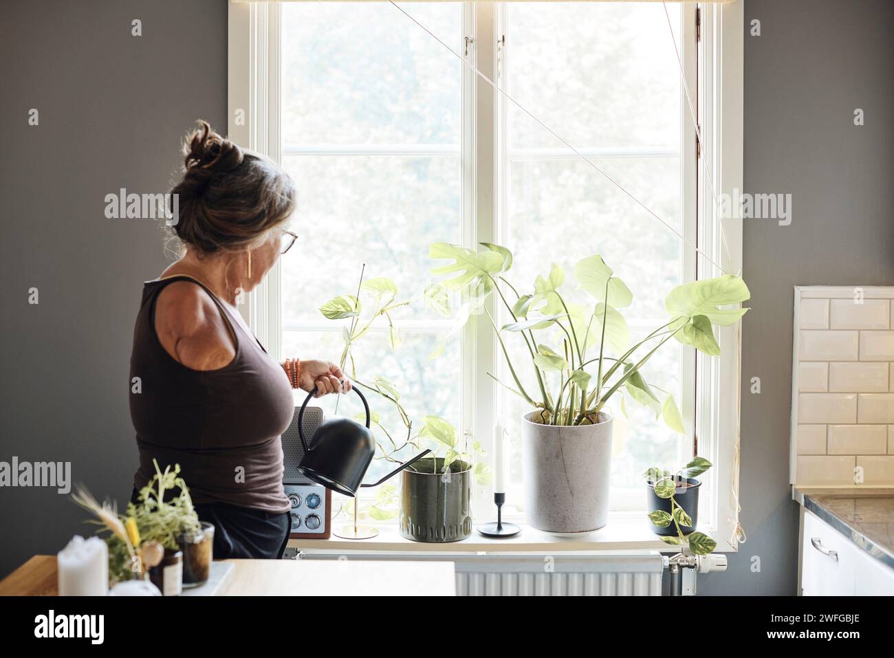 Reife Frau mit Behinderung, die Pflanzen auf der Fensterbank zu Hause tränkt Stockfoto