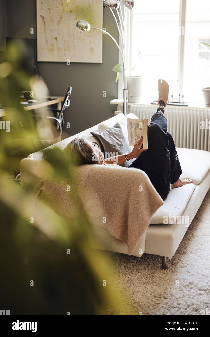 Reife Frau liest Buch, während sie sich zu Hause auf dem Sofa liegend Stockfoto