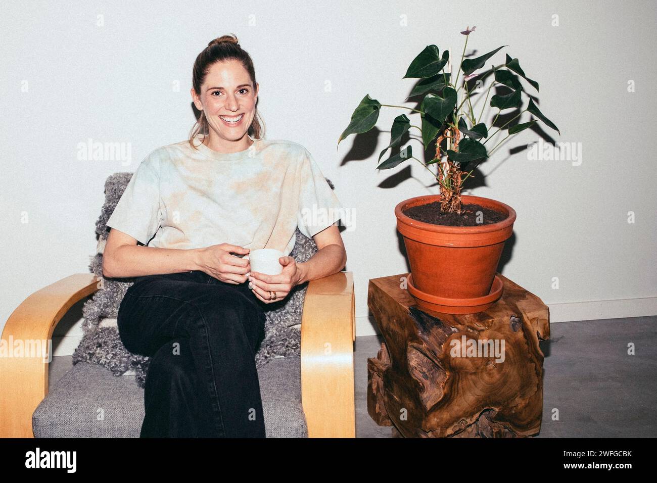 Porträt einer lächelnden Frau, die eine Teetasse hält, während sie zu Hause auf einem Stuhl neben einer Pflanze sitzt Stockfoto
