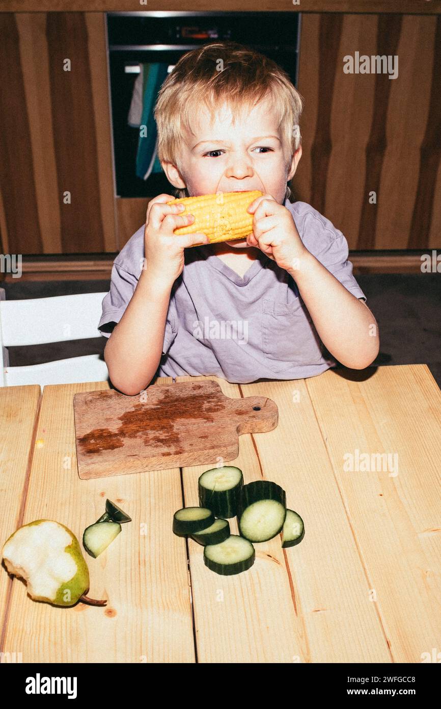 Hungriger Junge, der Mais beißt, während er zu Hause am Tisch sitzt Stockfoto