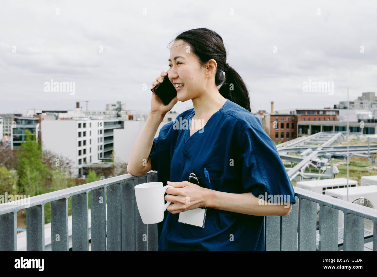 Lächelnde mittelerwachsene Krankenschwester, die eine Kaffeetasse hält, während sie mit dem Smartphone spricht Stockfoto