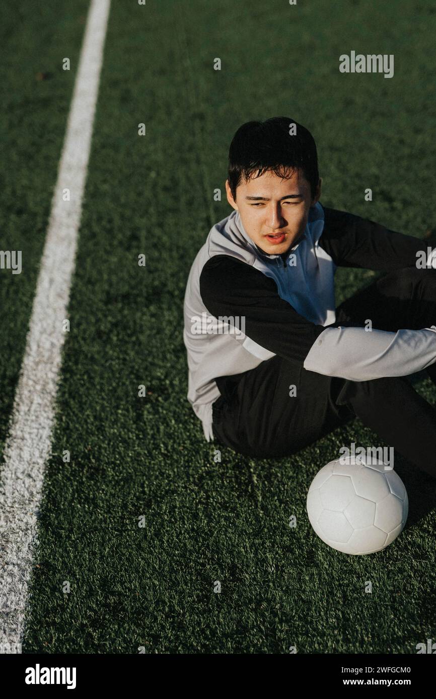 Hochwinkelansicht eines männlichen Athleten mit Fußball, während er auf dem Sportplatz sitzt Stockfoto