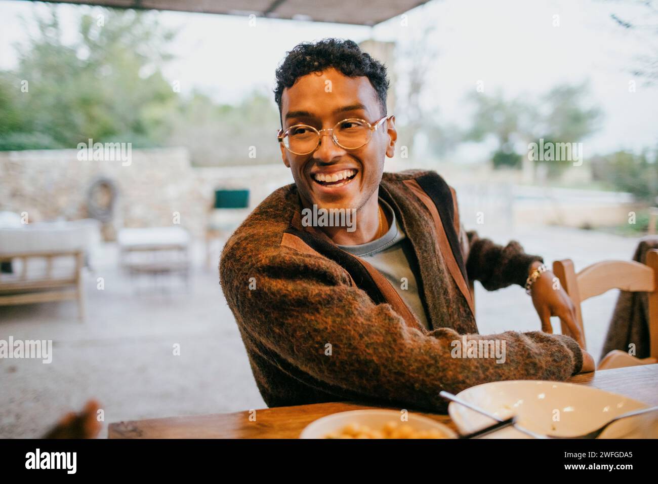 Glücklicher junger Mann mit Brille, der am Esstisch auf der Terrasse sitzt Stockfoto