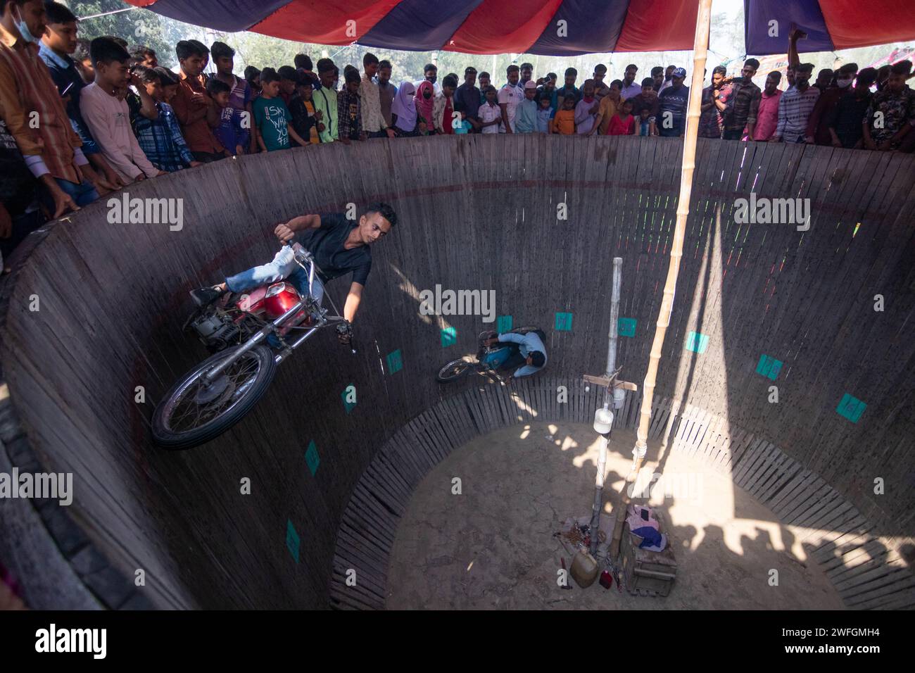 Dhaka, Khulna, Bangladesch. Januar 31, 2024. Waghalsige Autofahrer unterhalten die Zuschauer, indem sie gefährlich Motorräder auf einer vertikalen „Mauer des Todes“ in Khulna, Bangladesch, fahren. Bei Geschwindigkeiten von bis zu 80 km/h fahren die Künstler ohne Schutzausrüstung, damit ihre Fahrzeuge auf der 25 Fuß hohen Holzplanke balancieren können. Die Fahrer ziehen tödliche Stunts ab, um Zuschauer zu begeistern, um zusätzliches Geld zu verdienen, während sie senkrecht an einer Wand entlang fahren. Sie führen auf, um diejenigen zu unterhalten, die einen Eintritt von 20 Pence für die Unterhaltung bezahlt haben. Bis zu 100 Zuschauer können gleichzeitig dabei zusehen, wie sie Geld anbieten Stockfoto