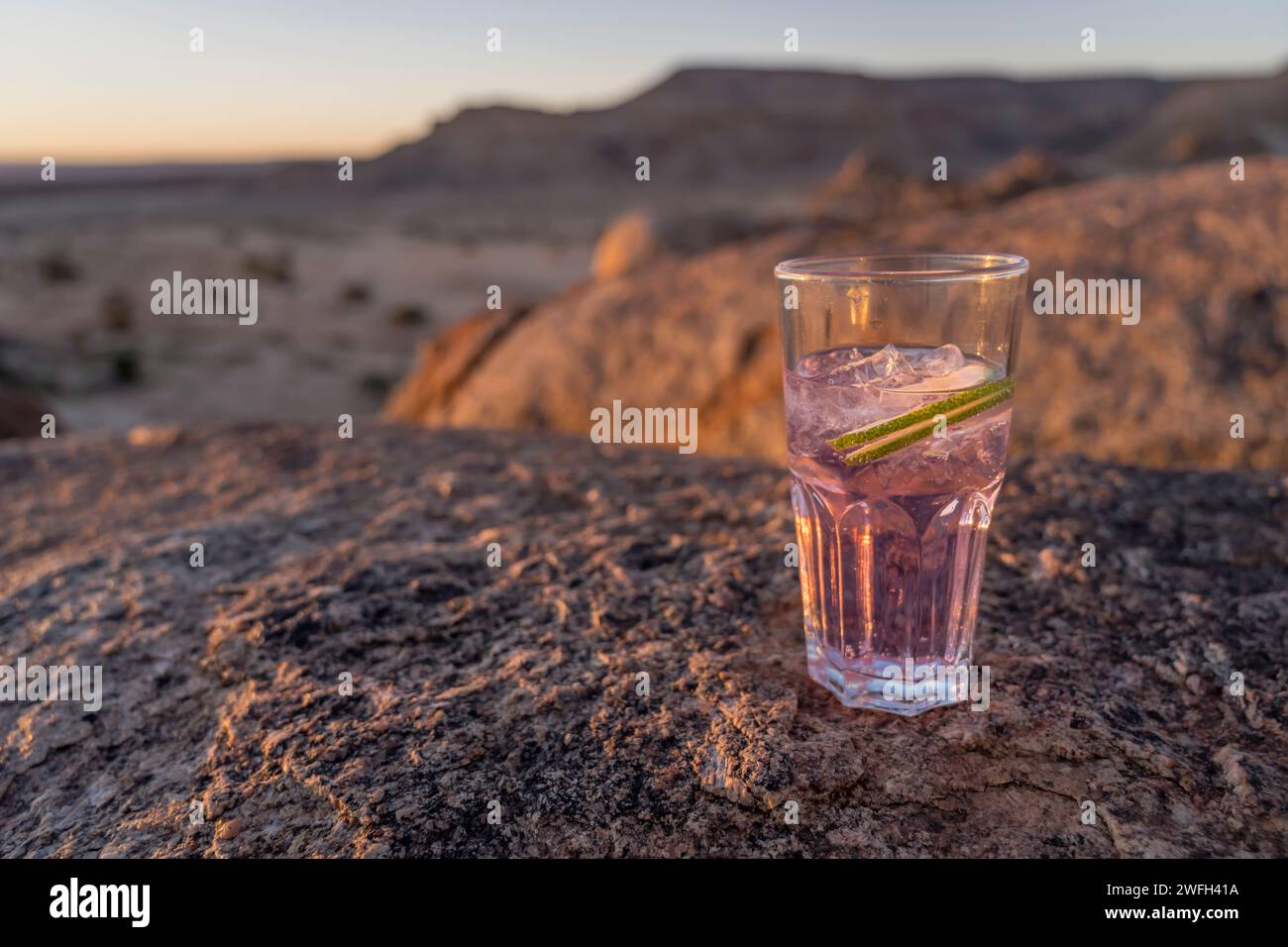Glas mit rosafarbenem Gin Tonic auf Doleritfelsen in der Wüste, aufgenommen im hellen Licht des späten Frühlings bei Sonnenuntergang in der Nähe von Hobas, Namibia, Afrika Stockfoto