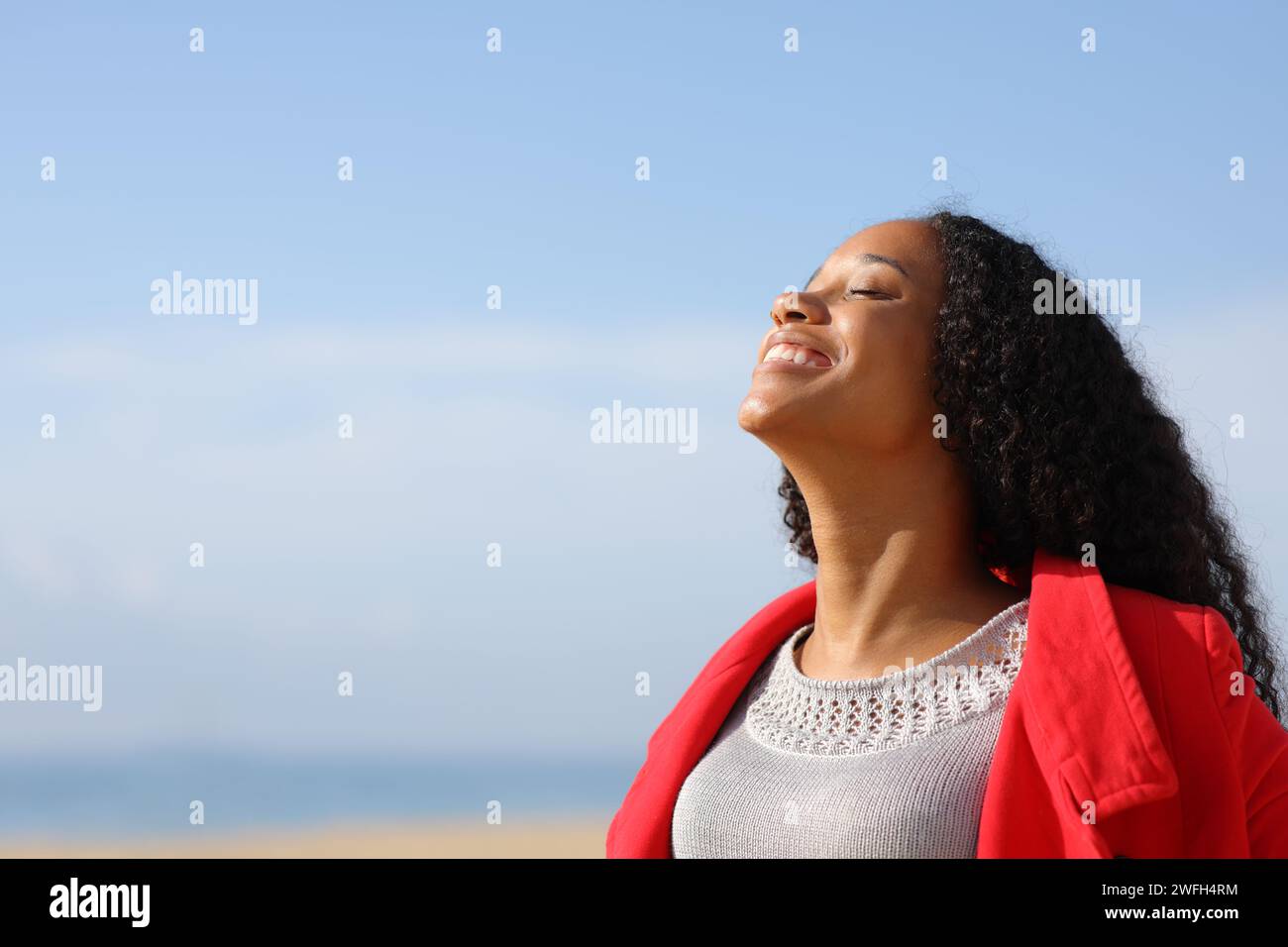 Glückliche schwarze Frau, die rote Jacke trägt und frische Luft am Strand atmet, im Winter an einem sonnigen Tag Stockfoto