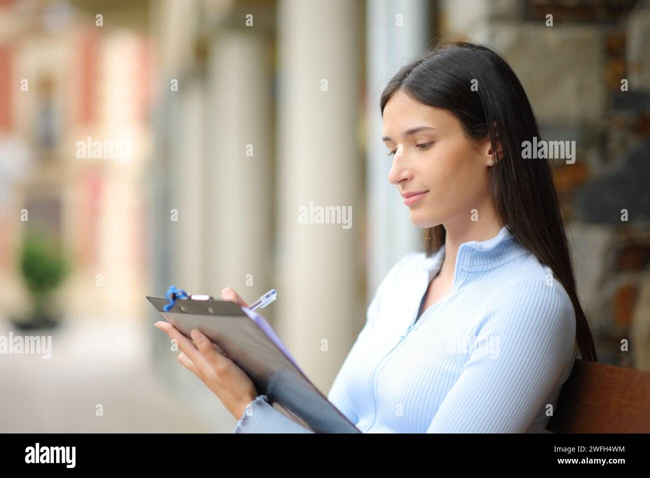 Ernsthafte Frau, die das Formular ausfüllt, sitzt auf einer Bank auf der Straße Stockfoto