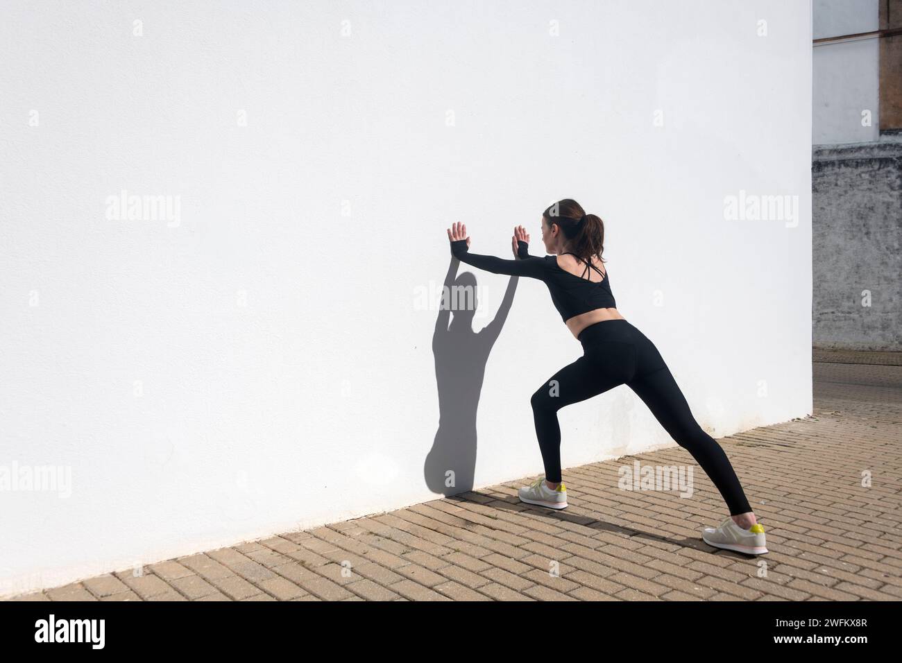 Sportliche Frau, die die Oberschenkelmuskulatur gegen eine weiße Wand dehnt Stockfoto