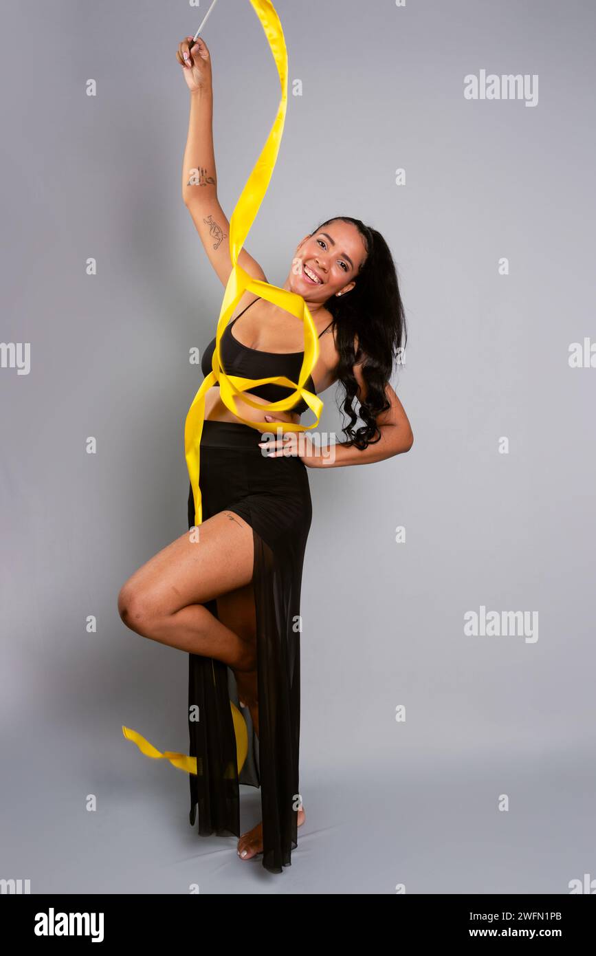 Schöne Turnerin, die akrobatische Bewegungen mit einem gelben Band macht. Isoliert auf grauem Hintergrund. Stockfoto