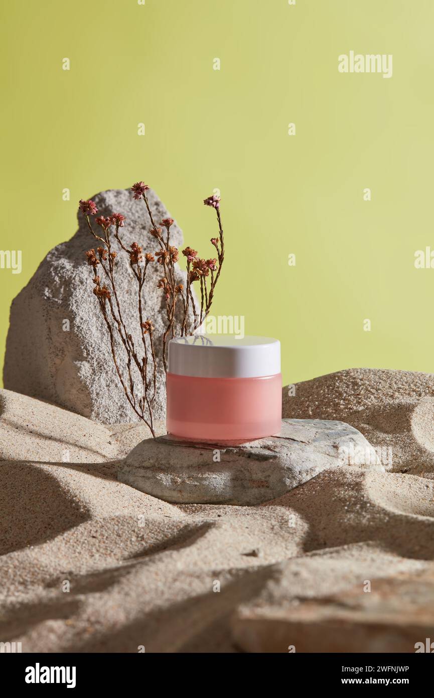 Ein Stein mit einem rosafarbenen Gefäß, verziert mit Blumen und ein paar Steinen auf dem Sand. Natürliche Hautpflege Beauty-Produktkonzept Stockfoto